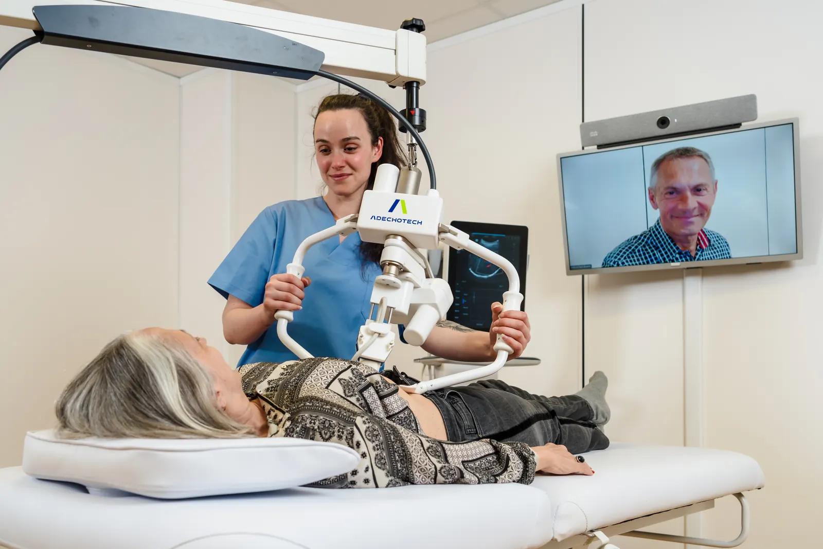 Abdominal examination with MELODY: a nurse holds the robotic arm on the abdomen of a patient lying down, the specialist is on the videoconference screen