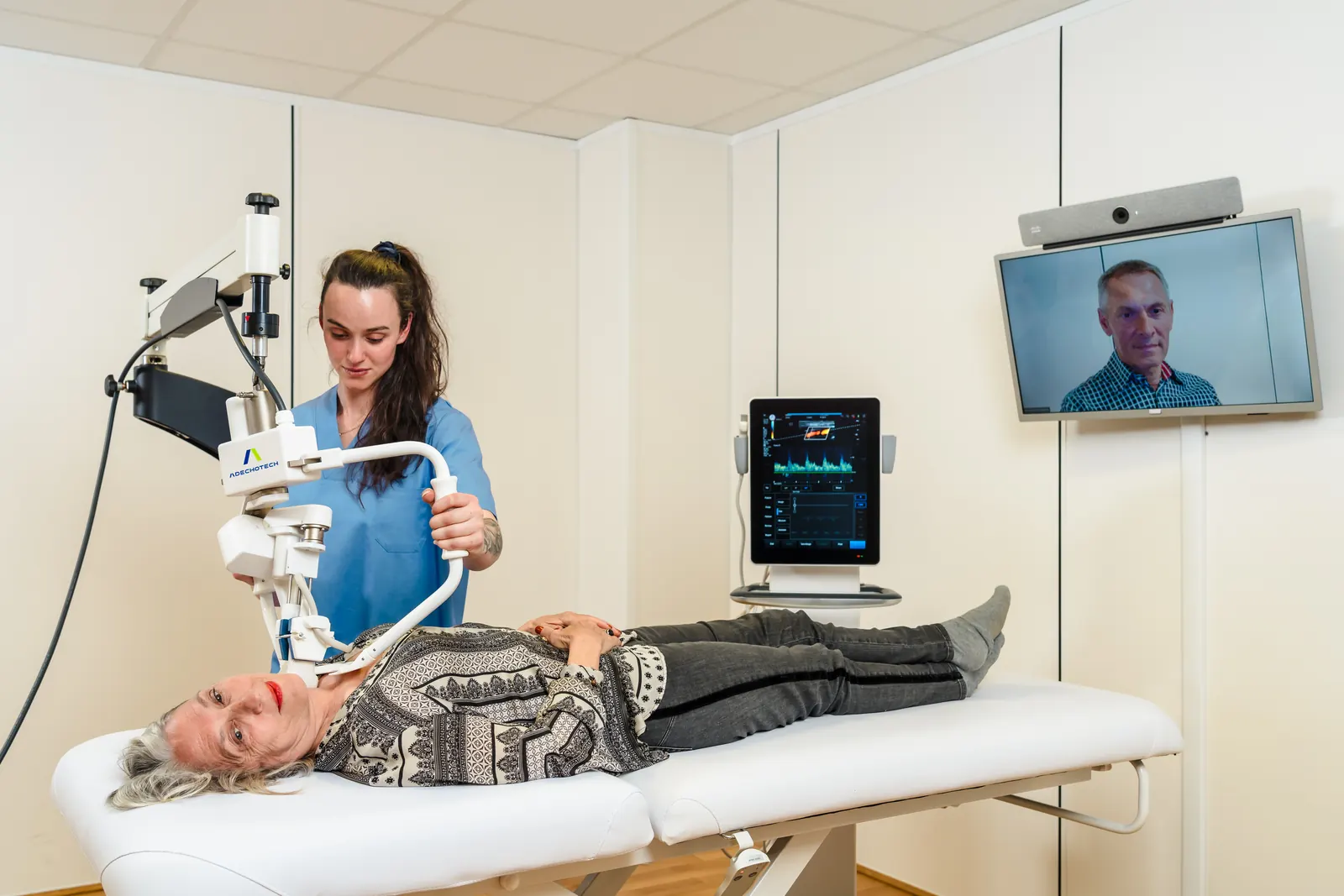 Examination room at the patient's site: patient lying down, nurse with the robotic arm, ultrasound system and videoconference screen