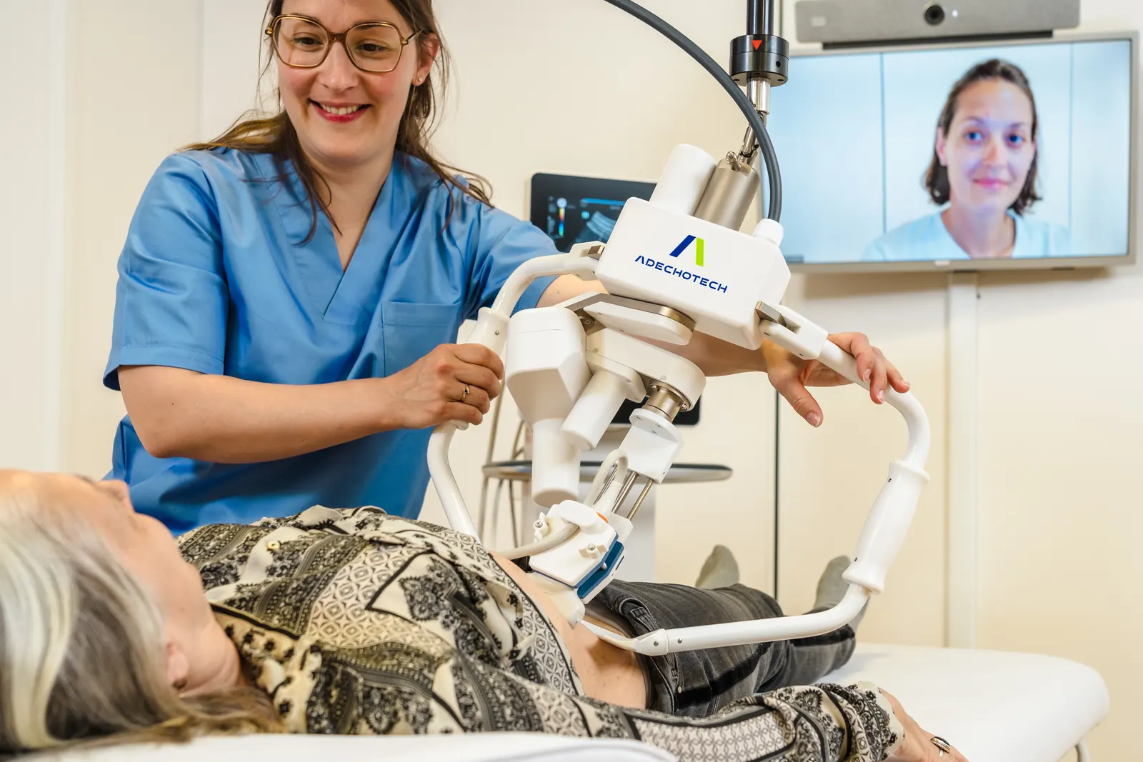 A smiling nurse guides the MELODY robotic arm during a patient's examination, the specialist on the videoconference screen
